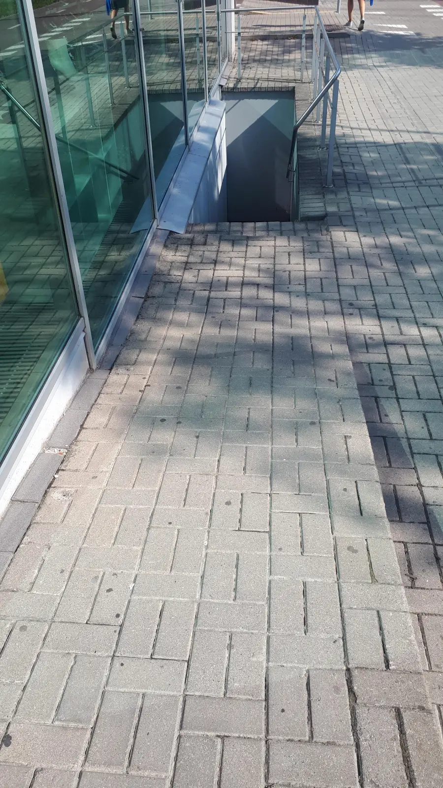 Sunlit sidewalk next to a glass building, with a stairway descending below ground level, surrounded by paved bricks; metal railings line the stairs.