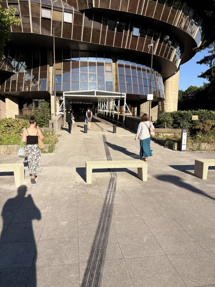 People walk toward a modern glass building entrance with benches and tactile paving guiding the way. Shadows and greenery are visible in the sunny outdoor scene.