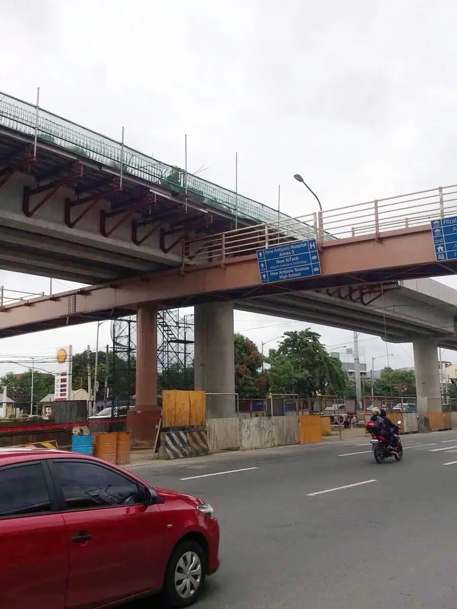 A red car and a motorbike pass beneath a partially constructed overpass and an existing overpass on a city street, with road signs and construction barriers visible.