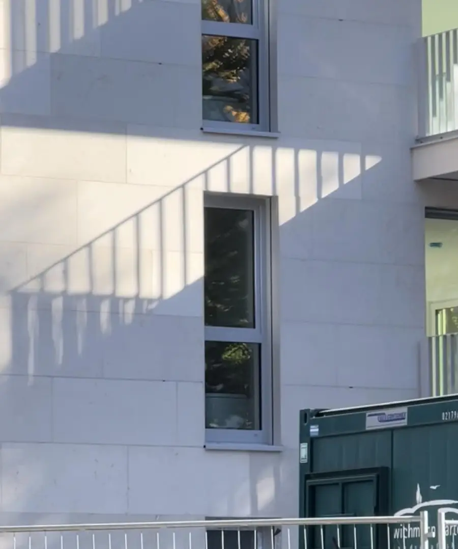 Sunlight casts diagonal shadows from a balcony railing onto a white building wall with two windows, creating a geometric pattern; a green container is visible at the bottom right corner.
