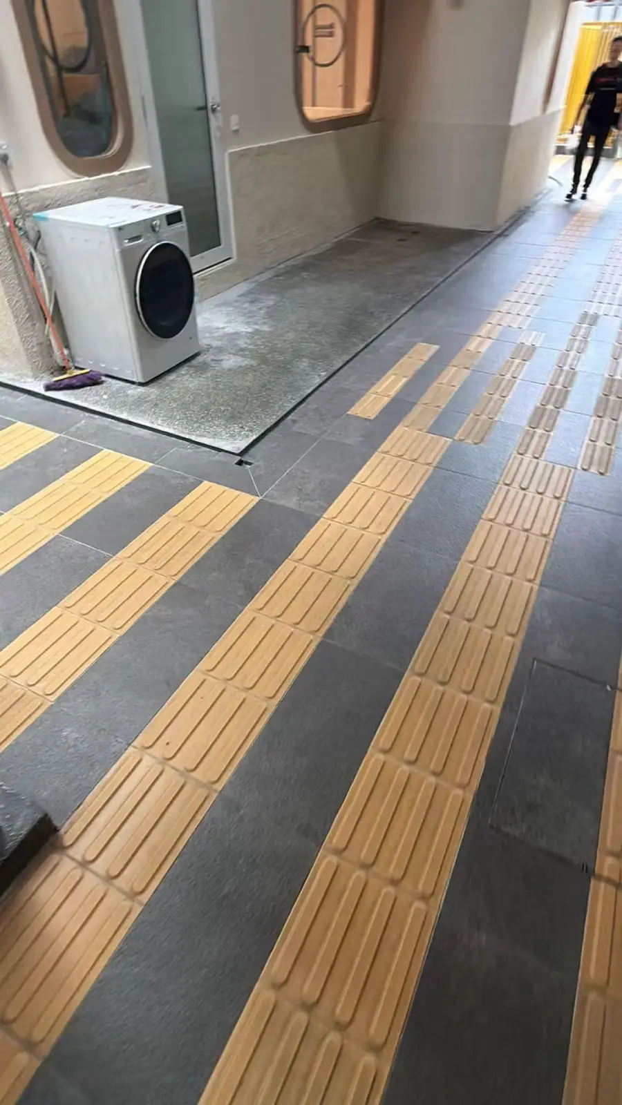 A washing machine blocks part of a tactile paving path in a hallway, making it difficult for visually impaired individuals to navigate. A person is visible walking in the background.