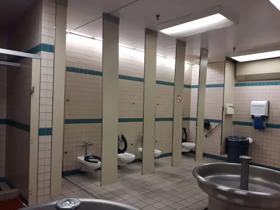 A public restroom with toilets separated by narrow vertical panels without doors, lacking privacy. The walls are tiled, and there are sinks and trash bins visible in the foreground.