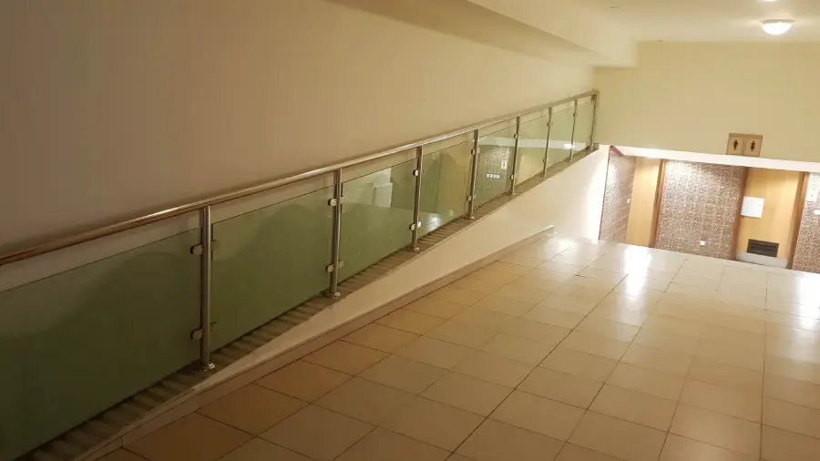 A tiled hallway with a glass and metal railing on the left side, leading down a gentle slope to a brightly lit area with patterned walls.
