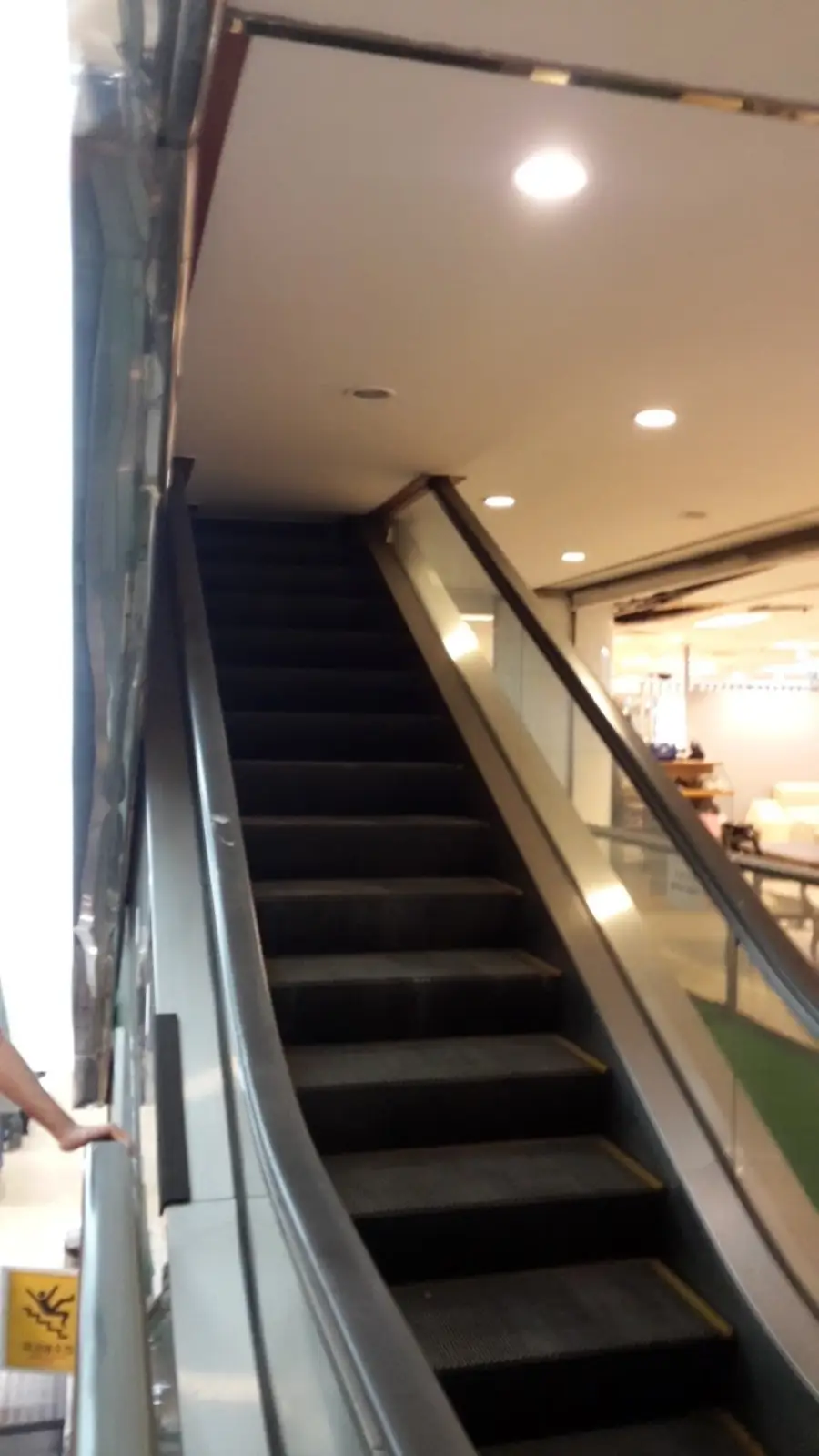 An escalator in a mall or store leads directly into a low ceiling, making it unusable. The area is lit with ceiling lights, and some store displays are visible to the right.