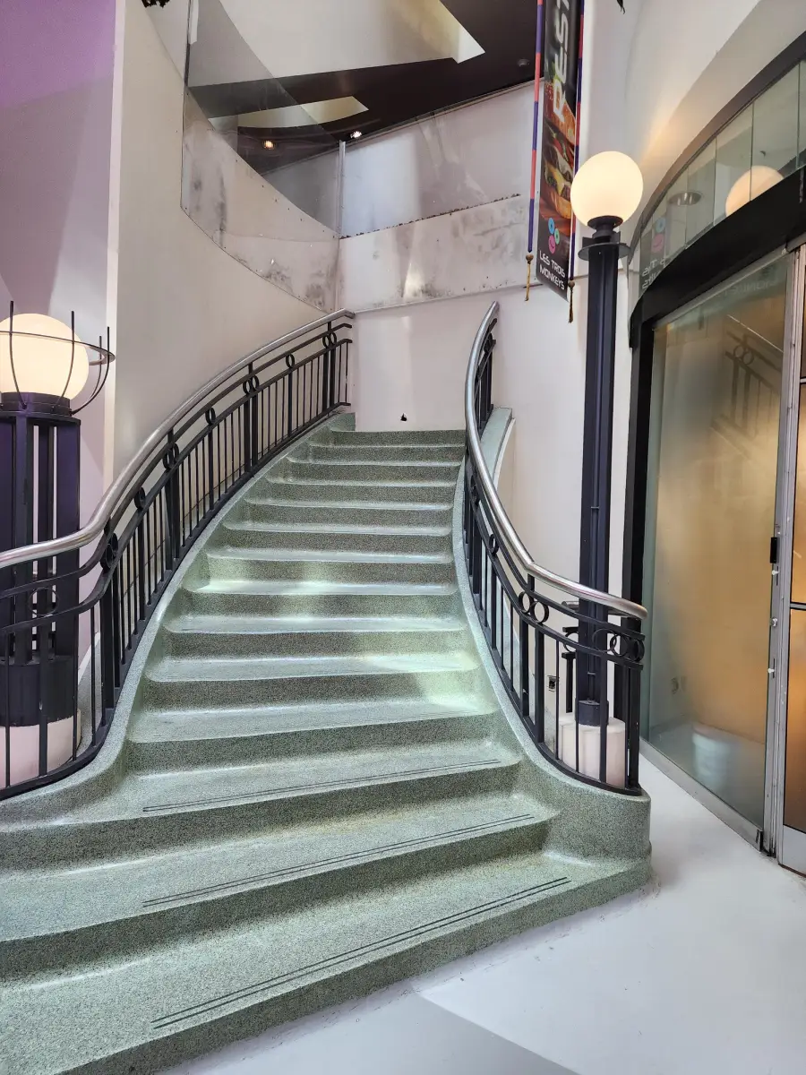 Curved indoor staircase with a metal railing, light-colored steps, glass panels above, and round lamps on both sides. There is a glass door on the right and banners hanging from the upper level.