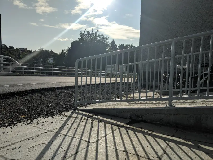 A metal railing casts long shadows on a ramp and pavement under bright sunlight, with part of a road, trees, and a building visible in the background.