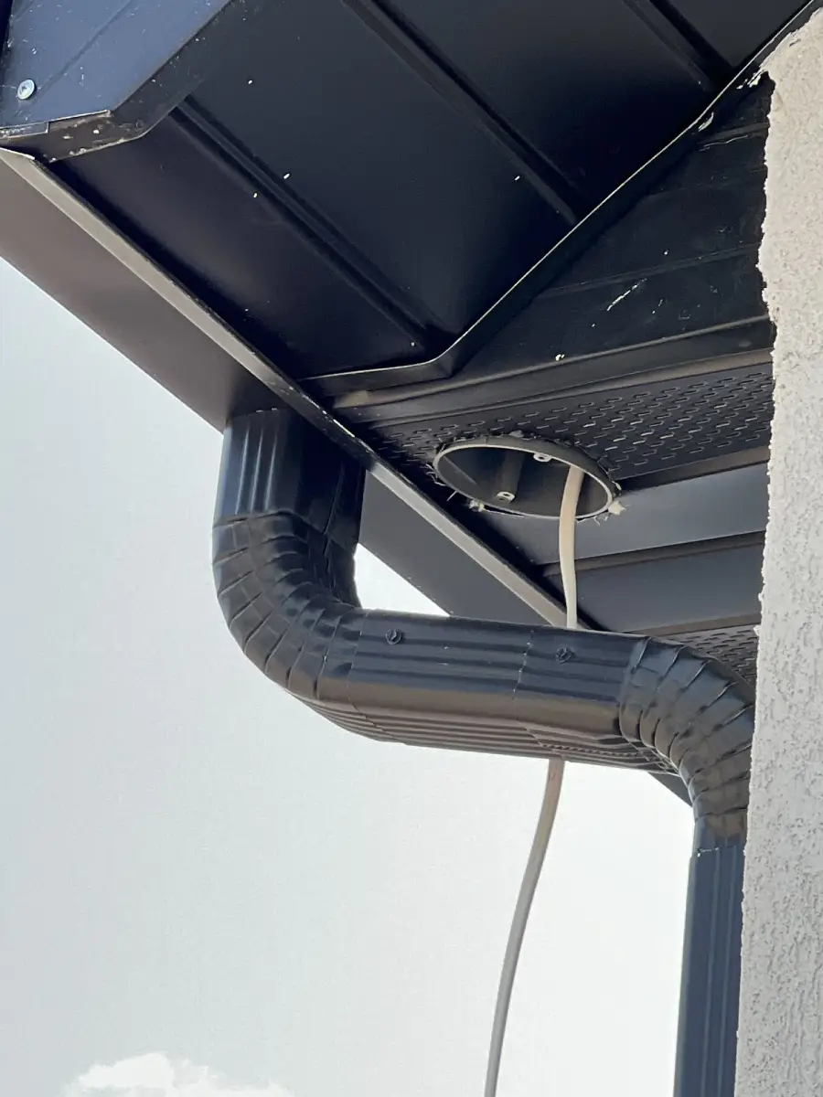 A black downspout bends awkwardly under a roof, with a white cable passing through a hole in the soffit panel, next to the gutter system, attached to the corner of a light-colored building.