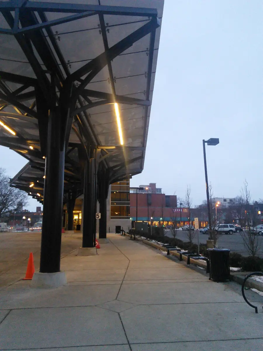 Covered bus terminal with numbered sign, orange cones, and sidewalk at dusk. Buildings, cars, bare trees, and streetlights are visible in the background.