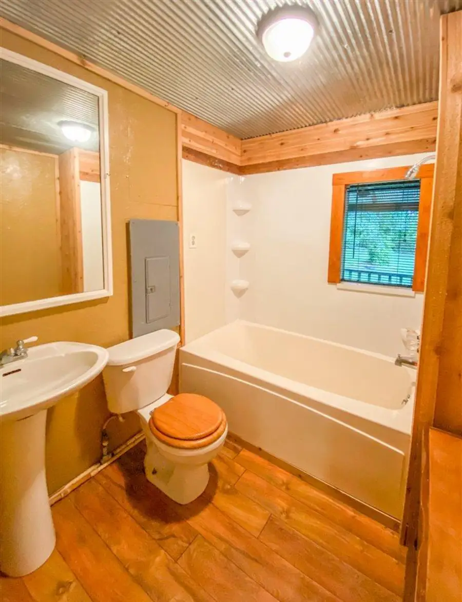 Small bathroom with wood floors, a pedestal sink, toilet with a wooden seat, bathtub, mirror, metal ceiling, and a window with a blind. Exposed wood trim adds a rustic feel.