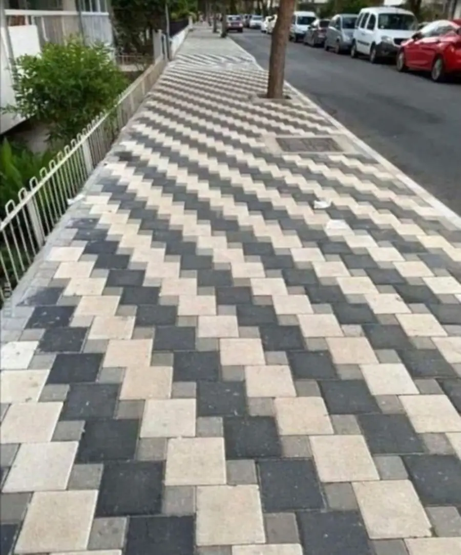 A sidewalk with black, gray, and white tiles arranged in a zigzag pattern, creating an optical illusion of a wavy, uneven surface next to a street with parked cars.