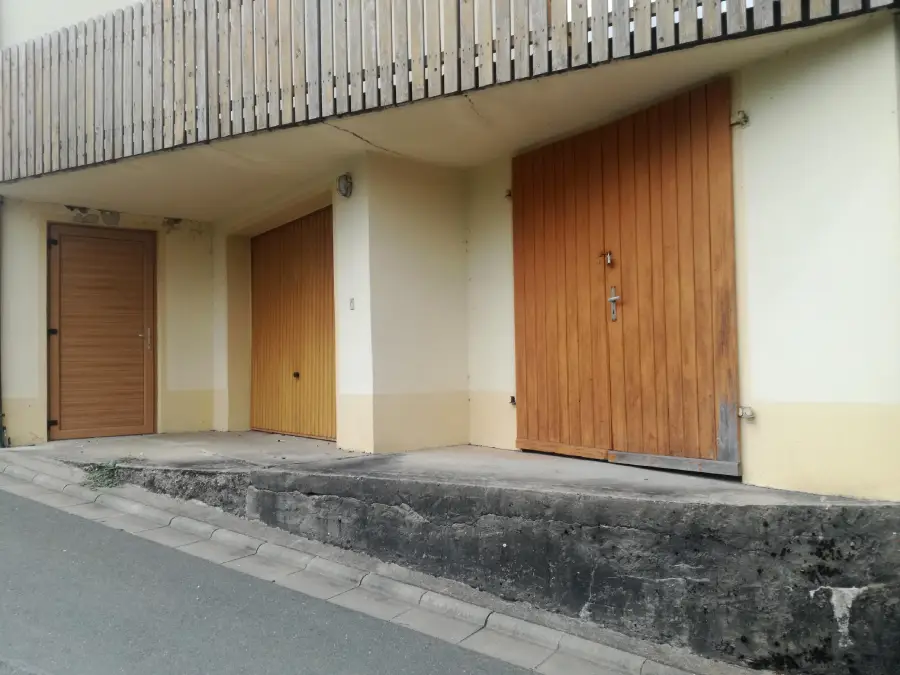 Three wooden doors of different sizes in a pale yellow building, set above a concrete platform next to a sloped road with a curb. A wooden fence runs above the doors.