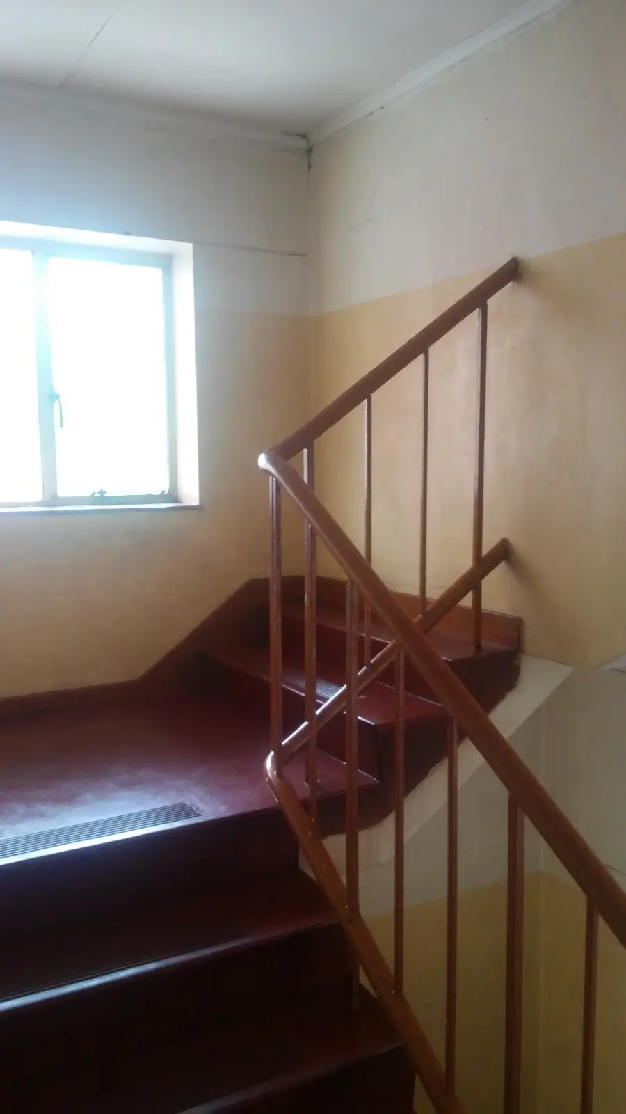 A stairwell with brown steps and railing, yellow and white walls, and a window letting in daylight.