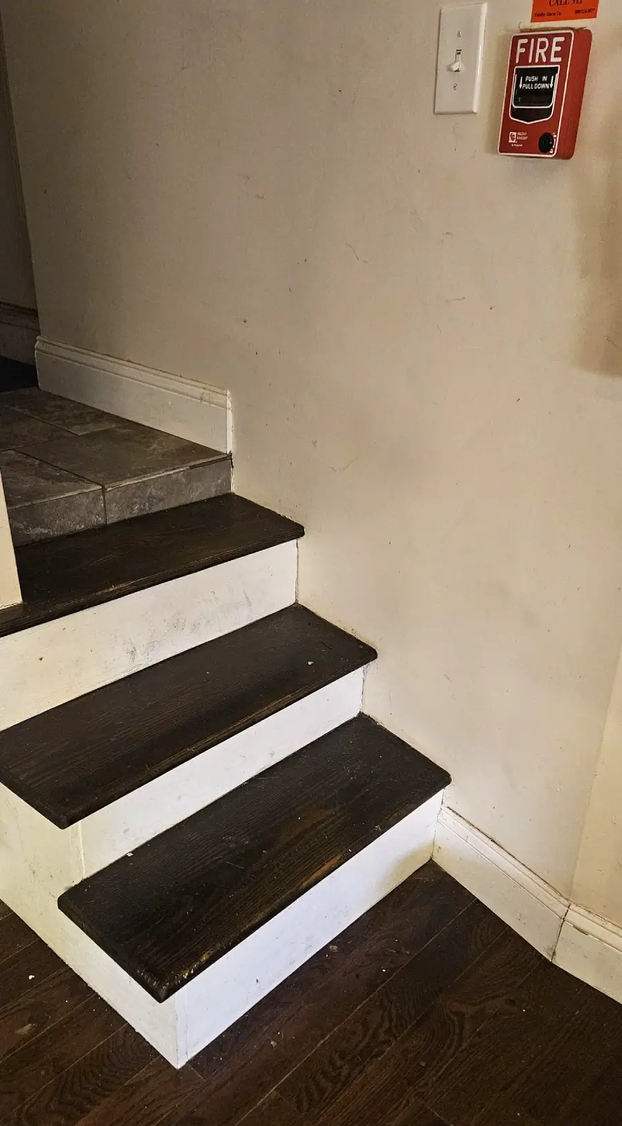 Three wooden steps lead up to a tiled floor. A red fire alarm pull station and a white electrical outlet are mounted on the beige wall beside the stairs.