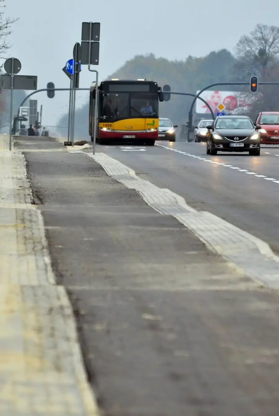 A city street with a bus and several cars driving, traffic lights ahead, and a sidewalk running alongside the road on a cloudy day.