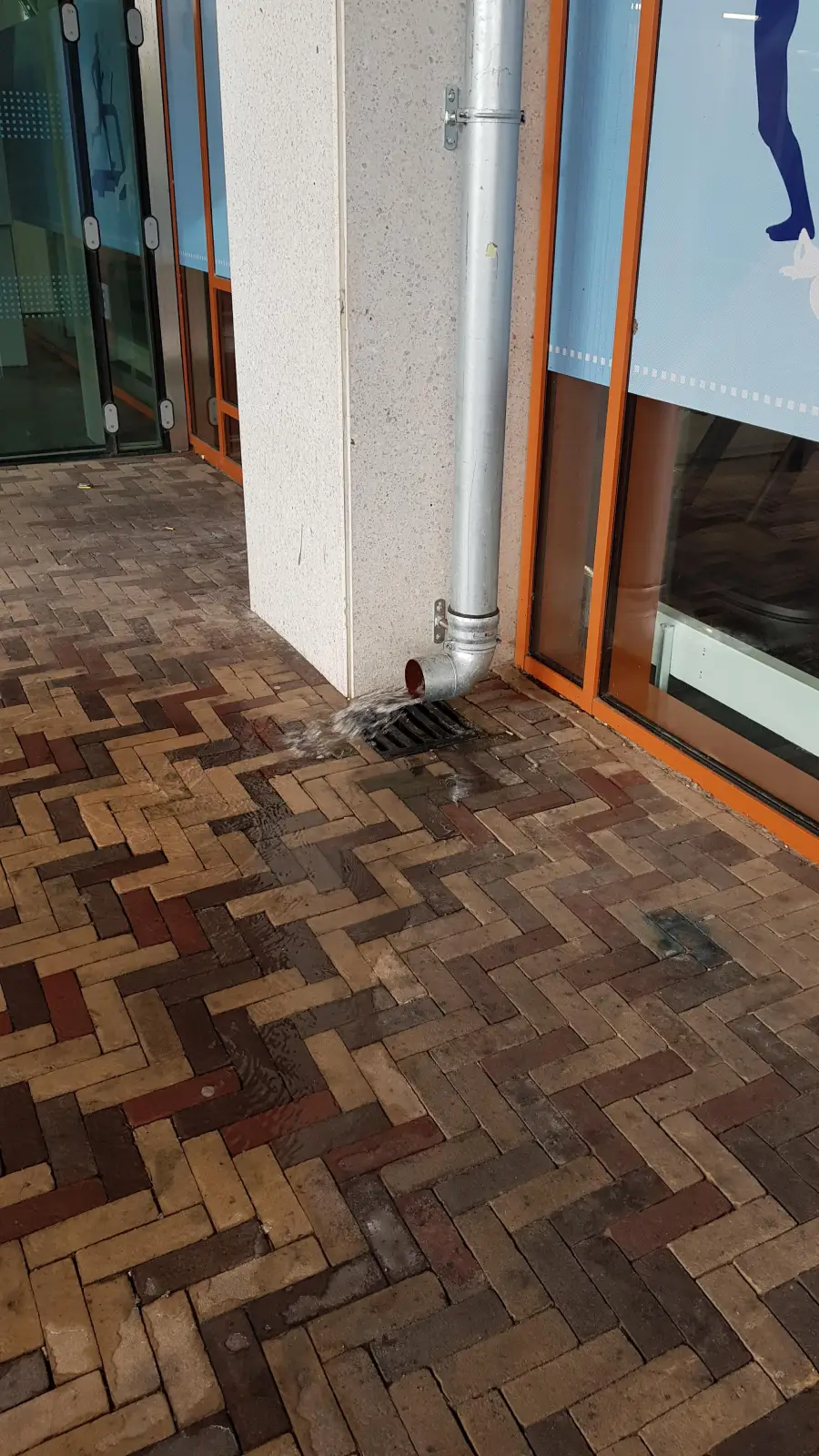 A downspout drains water onto a brick sidewalk, causing a wet patch near a building with glass windows and orange trim.