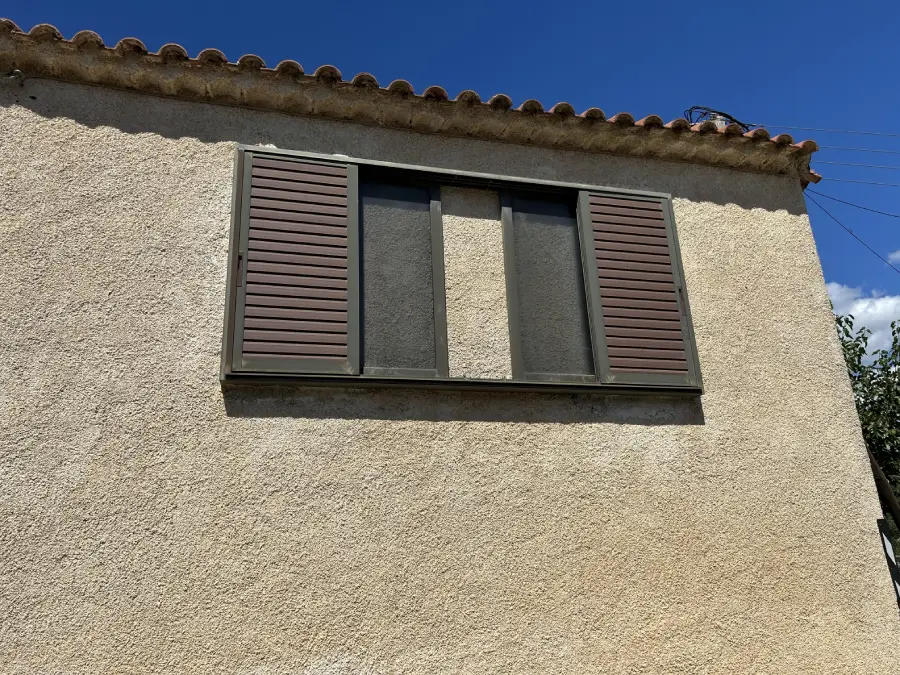 A beige stucco wall with a window featuring two closed brown shutters on each side and a covered center pane, set against a blue sky.