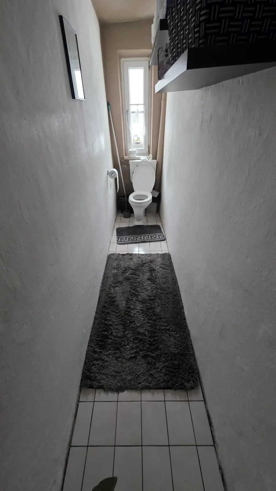 A narrow bathroom with white walls, a gray rug, and a toilet at the far end under a small window. There is a framed picture on the left wall and a shelf with a basket on the right.