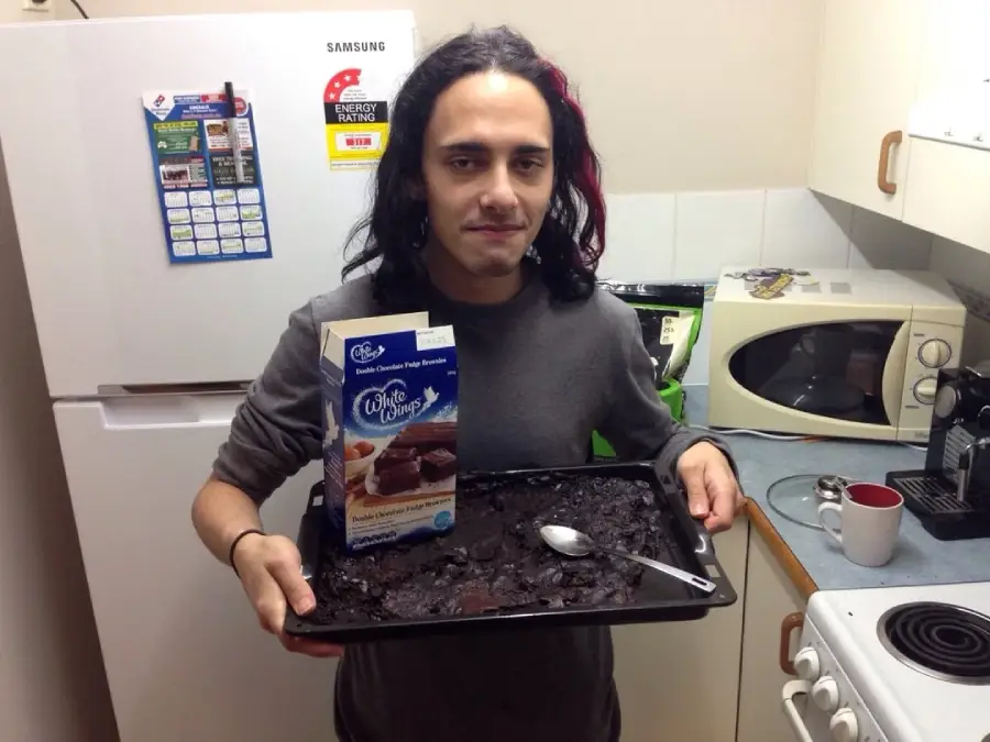 A person with long dark hair holds a tray of brownies and a boxed mix in a kitchen. The brownies look burnt or undercooked, with a spoon in the tray. Kitchen appliances and mugs are visible in the background.