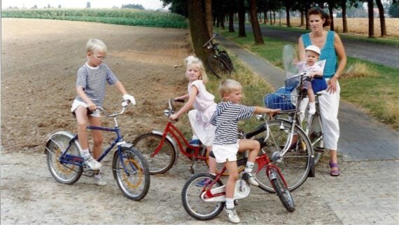 A woman stands with a baby in a bike seat while three young children ride small bikes on a rural path beside a field. All wear summer clothes; two boys wear striped shirts. Trees and another bike are in the background.