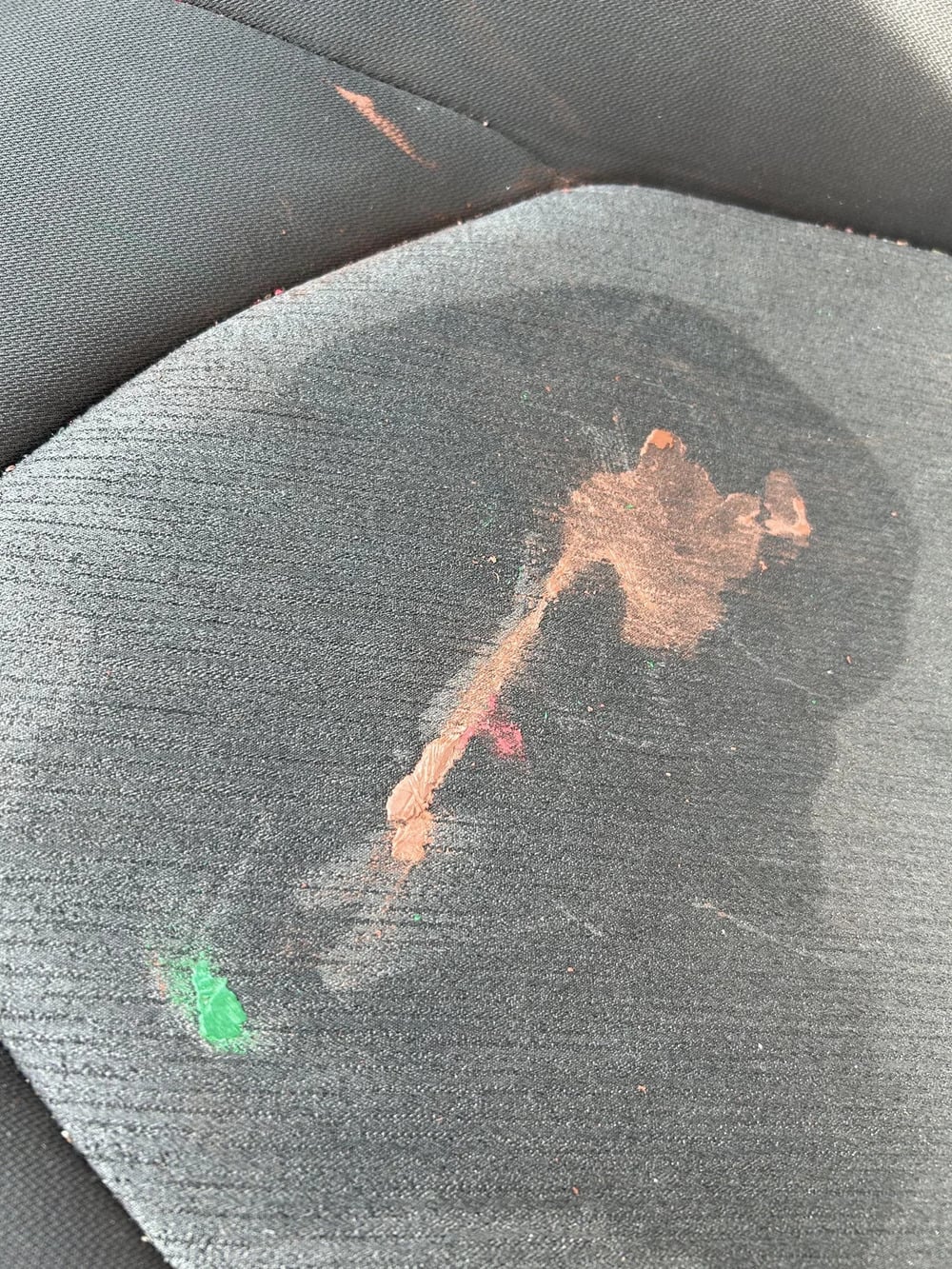 A close-up of a car seat with a large brownish stain and small green and red spots on the black fabric upholstery.