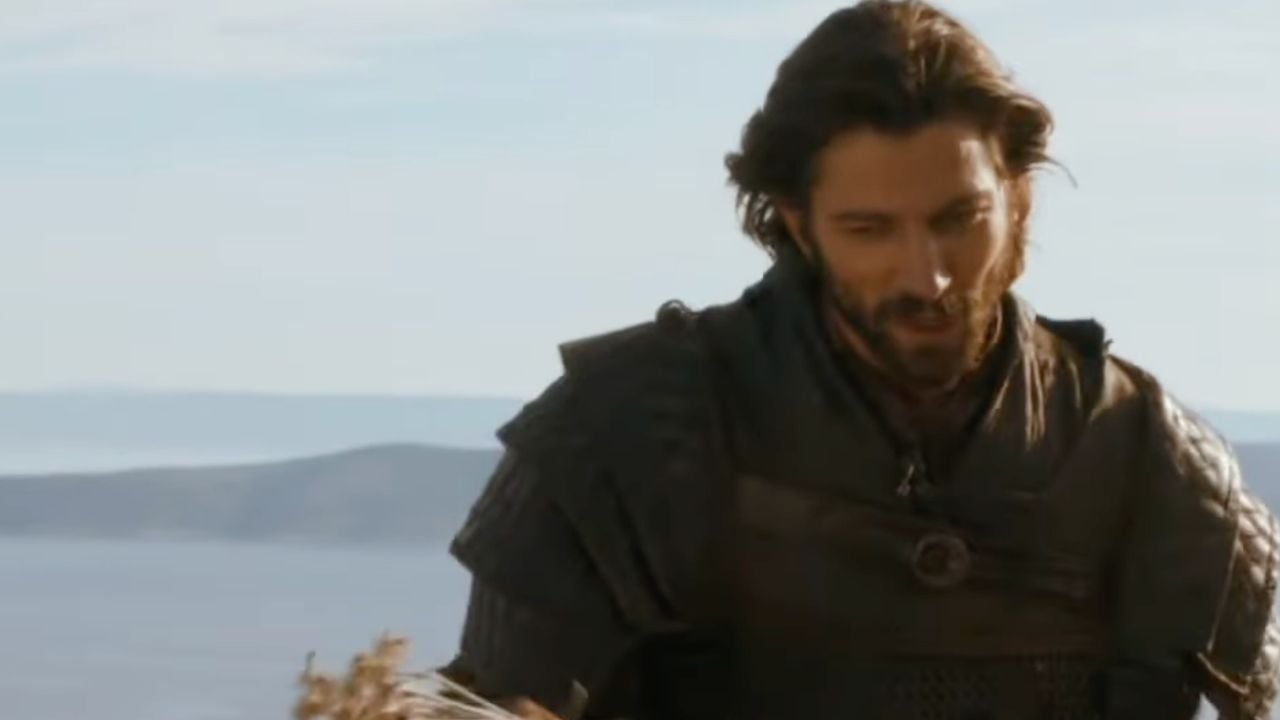A man in dark medieval armor stands outdoors with mountains and a body of water in the background under a clear sky.
