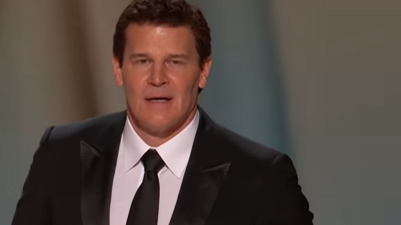A man in a black tuxedo and black tie speaks on stage against a blurred background.