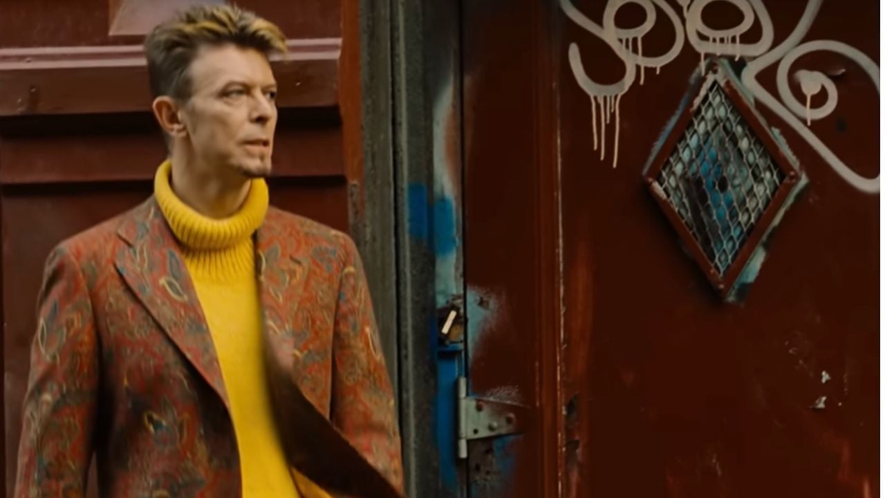 A person with short, styled hair in a yellow turtleneck and patterned jacket stands near a red door with graffiti and a diamond-shaped window.
