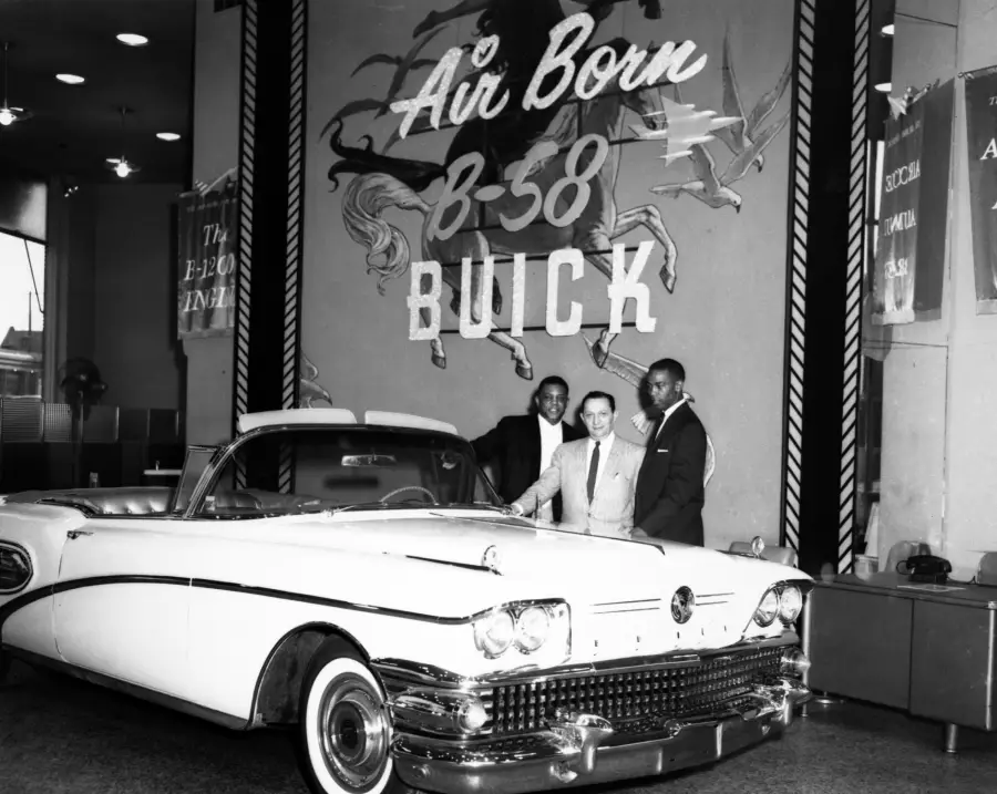 Three men stand beside a classic Buick convertible in a showroom. Behind them is a large sign that reads “Air Born B-58 BUICK.” The atmosphere is bright and formal.