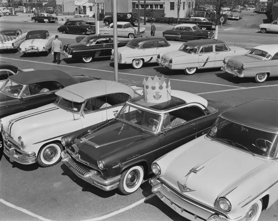 Vintage black-and-white photo of classic cars parked in a lot, with one car featuring a large Burger King crown on its roof. People and more cars are visible in the background.