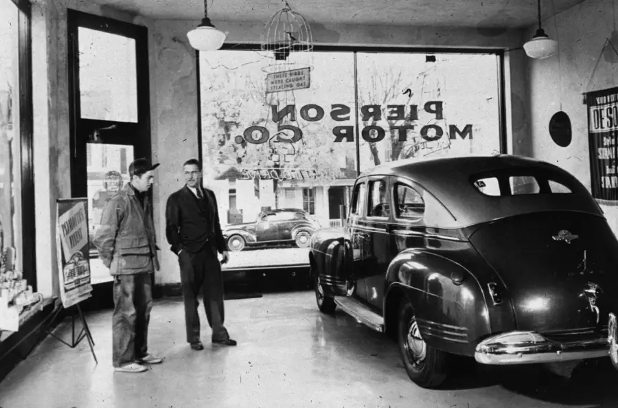 Two men stand inside a car showroom, looking at a vintage automobile. Large windows reveal a street and parked cars outside. A sign on the window reads “PIERSON MOTOR CO.”