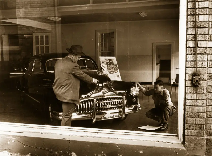 Two men examine a vintage car inside a showroom; one stands with a hand on the hood while the other crouches, measuring or inspecting the front. A poster and bare-walled interior are visible in the background, seen through a window.