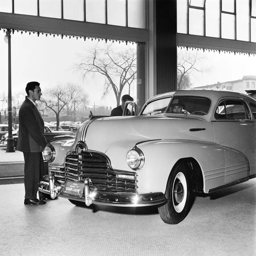 A man in a suit stands next to a classic car inside a showroom with large windows. Trees and parked cars are visible outside. The scene appears to be from the mid-20th century.