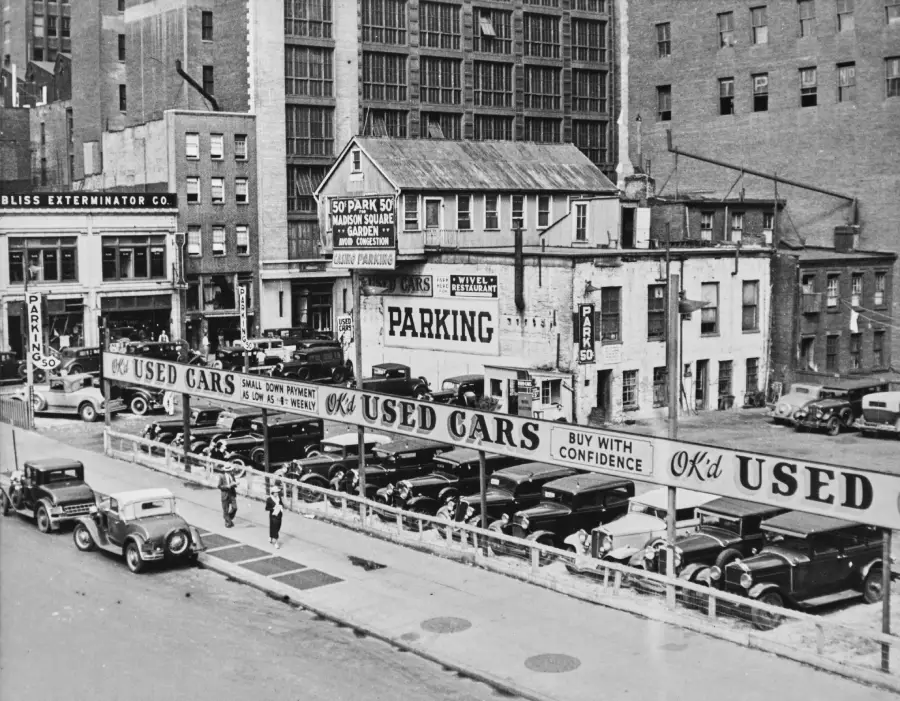 Black and white photo of a 1930s city street with a used car lot full of vintage cars, tall buildings in the background, street signs, and people walking on the sidewalk.