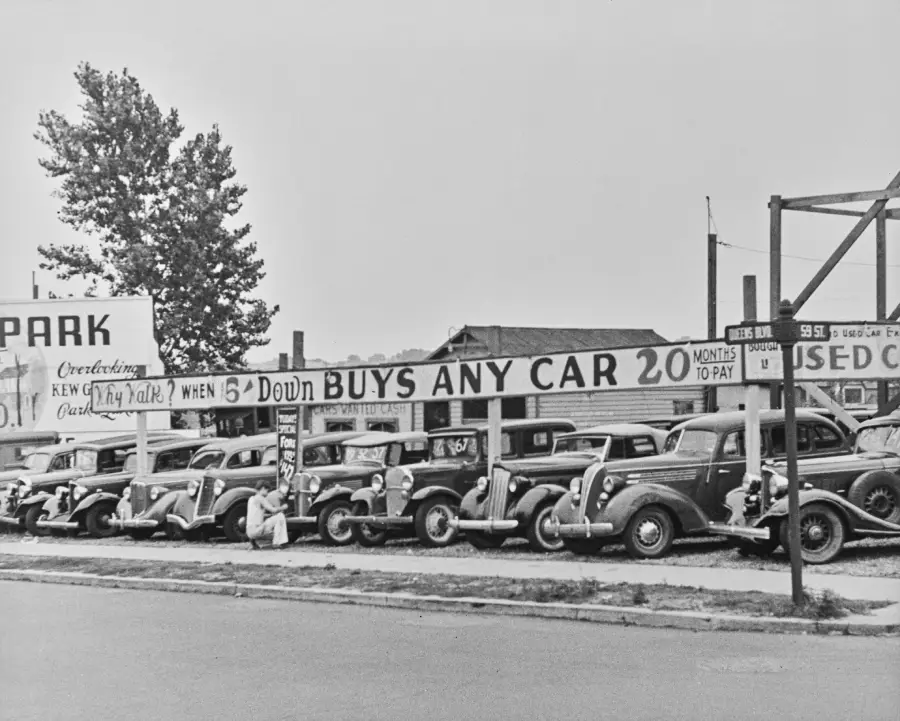 A row of vintage cars is lined up at a used car lot with a large sign reading “$5 down buys any car, 20 months to pay.” Several people are inspecting the vehicles.