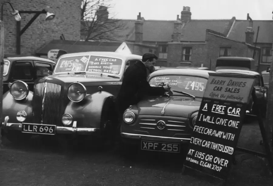 A black-and-white photo of a man writing on a car at a used car lot. Signs advertise car prices and a special offer: a free car with every car purchased at £195 or over. Other parked cars and buildings are visible in the background.