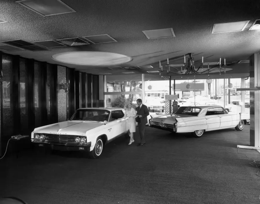A man and woman stand in a car showroom with two vintage cars, one parked on each side. The room has large windows, a chandelier, and a circular ceiling feature.