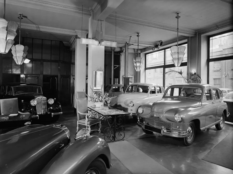 A black and white photo of a vintage car showroom, featuring several classic cars parked inside, decorative ceiling lights, large windows, and a table with a vase of flowers in the center of the room.