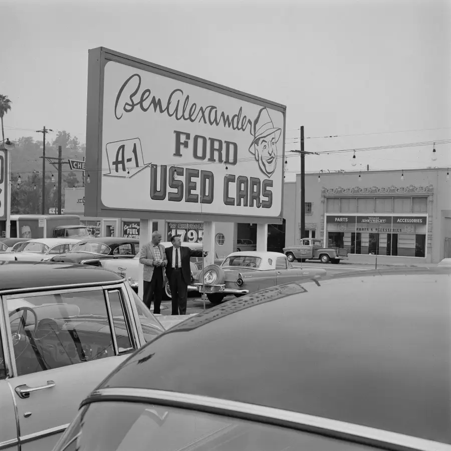 Black-and-white photo of a used car lot with a large sign reading “Ben Alexander Ford A-1 Used Cars.” Several cars are parked, and two men in suits are walking near the sign.