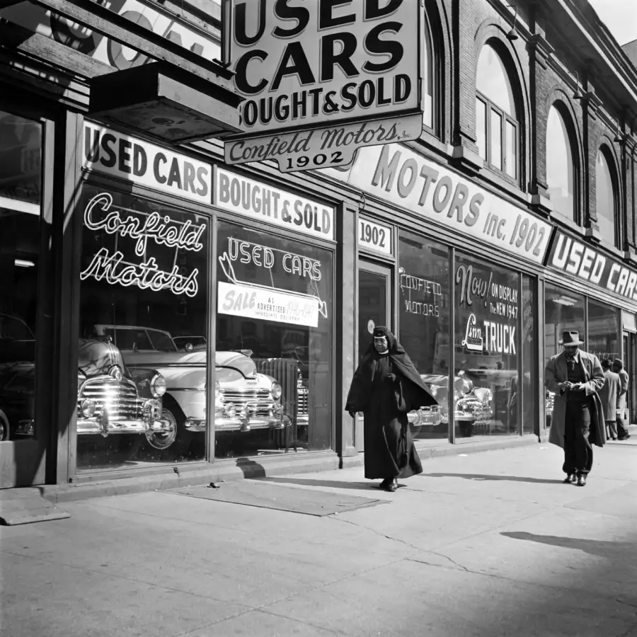 Black-and-white photo of a 1940s or 1950s used car dealership with neon signs in the window and vintage cars inside. Two people walk on the sidewalk in front of the storefront.