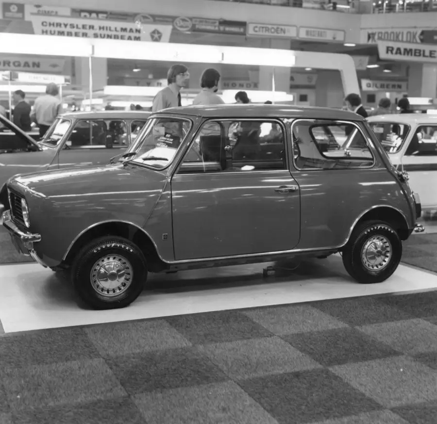 A classic compact car is displayed indoors at an auto show, surrounded by other vintage vehicles and exhibition signs. Several people are looking at the cars in the background.
