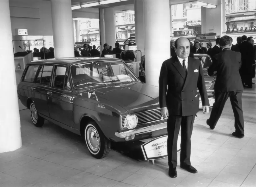 A man in a suit stands in front of a classic station wagon car displayed indoors, with other people and cars in the background. The scene appears to be from a vintage showroom or auto exhibition.