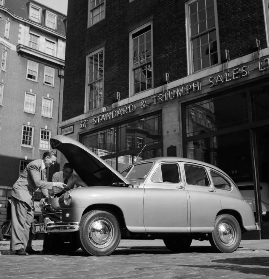 Two people inspect the engine of a vintage car with its hood open, parked outside a building labeled “STANDARD & TRIUMPH SALES LTD” in an urban setting.
