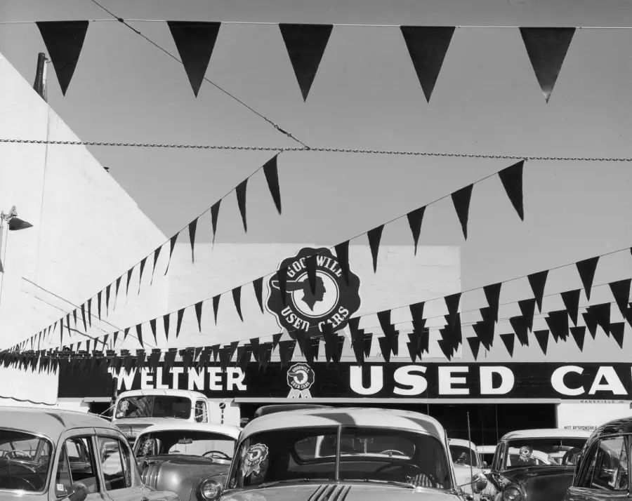Black and white photo of a used car lot with pennant flags overhead. Vintage cars are parked, and a sign reads “Weltner Used Cars” with a circular “Joe Wil” logo above.