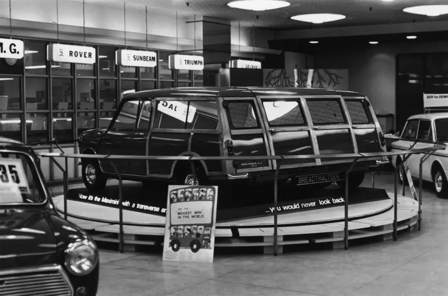 Black-and-white photo of a vintage station wagon displayed on a rotating platform at an indoor auto show, with informational signs and other cars visible nearby.