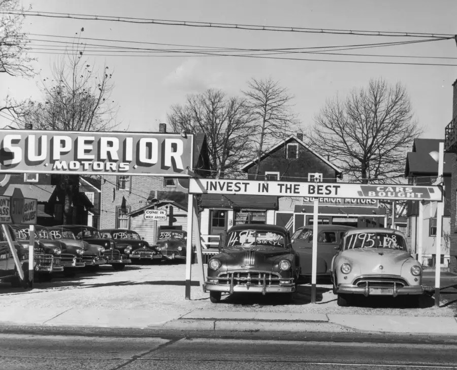 Black and white photo of a vintage car dealership with a “Superior Motors” sign, displaying classic cars for sale. Houses and leafless trees are visible in the background.