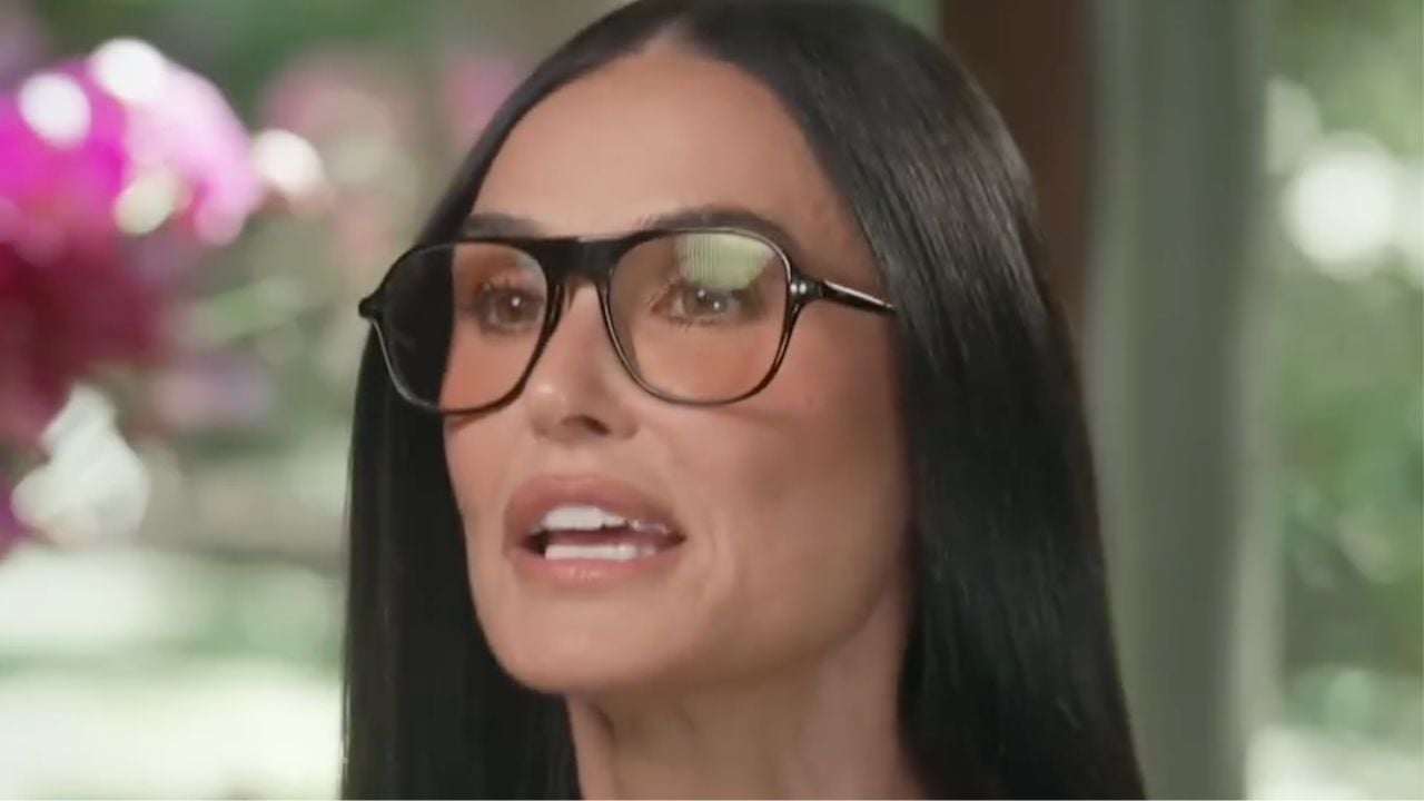 A woman with long straight dark hair, wearing large square eyeglasses, is speaking indoors with a blurred background of pink flowers and greenery.