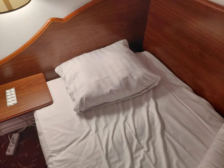 A neatly made single bed with a white pillow and sheets, wooden headboard, a small bedside table with a remote control, and a power strip on the floor beside the bed.