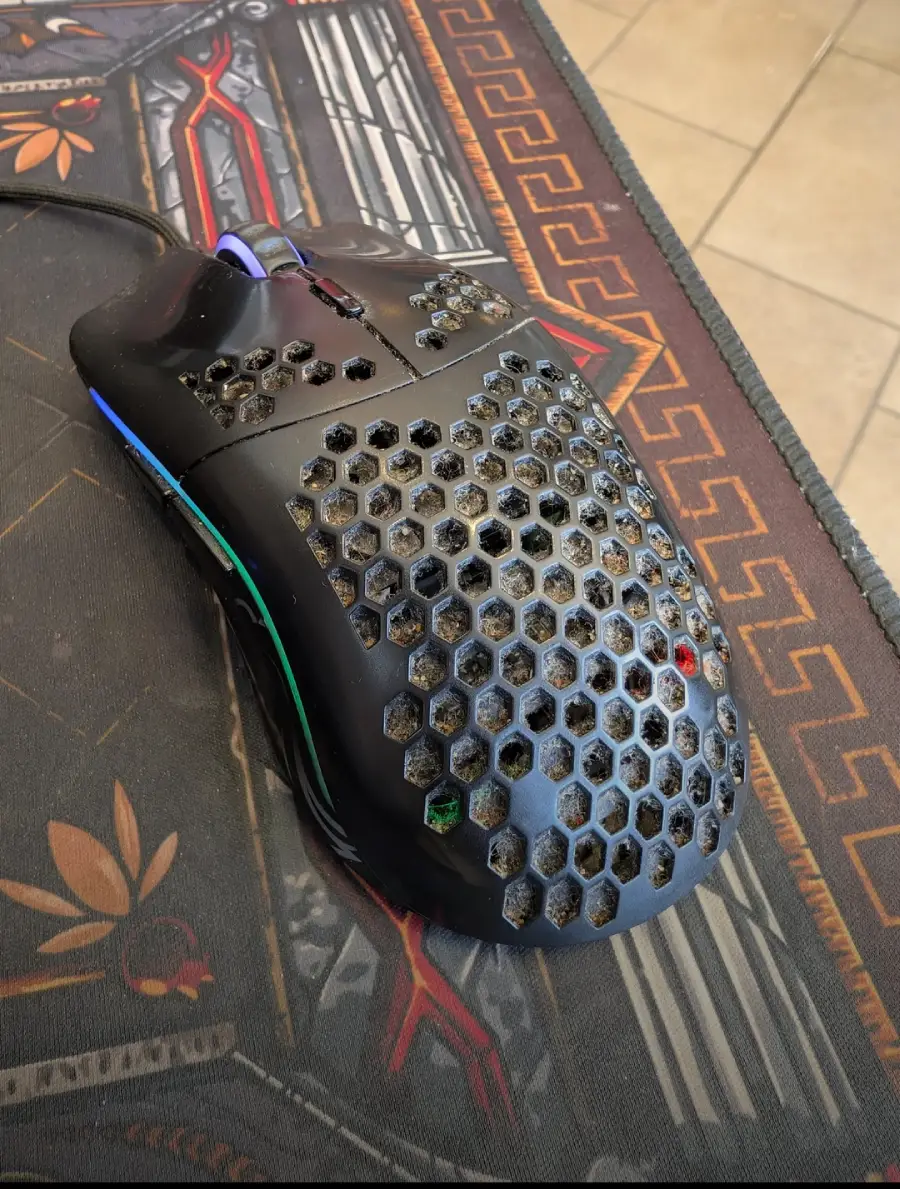 A black gaming mouse with a honeycomb design has visible dirt and debris inside the holes, placed on a decorative mouse pad with geometric and floral patterns.