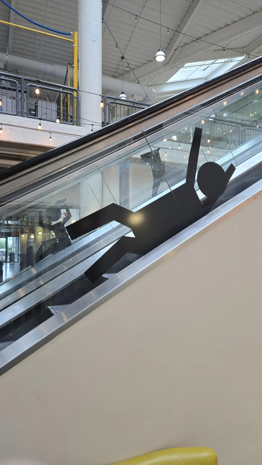 A black silhouette of a person appears to be falling on the side of an escalator in a modern indoor space with lights and railings in the background.