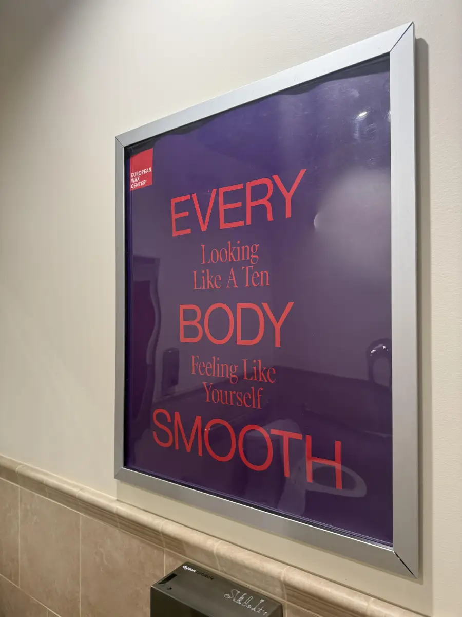 A framed poster with purple background and red text reads: “EVERY Looking Like A Ten BODY Feeling Like Yourself SMOOTH.” A bathroom sink and soap dispenser are visible in the reflection.